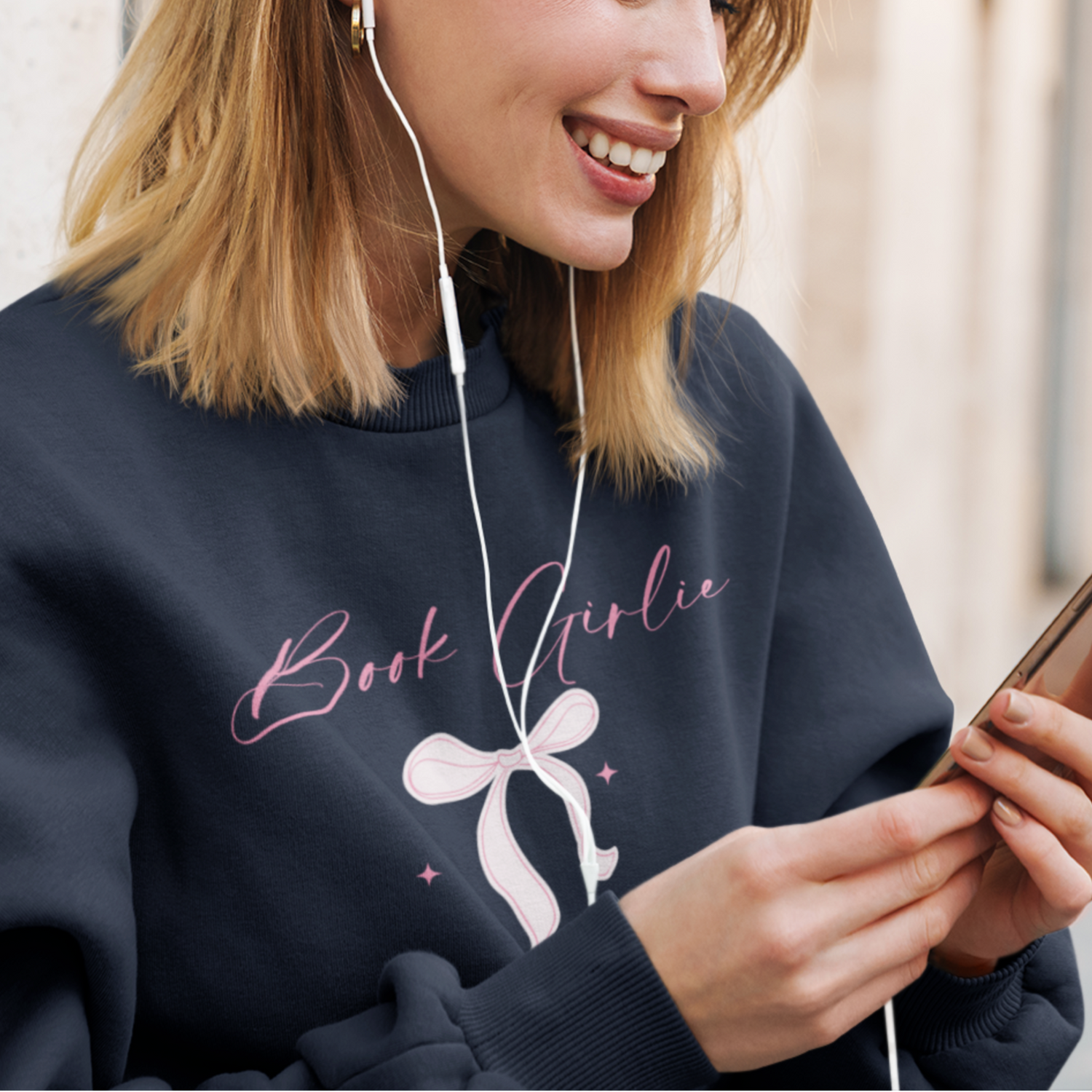 Woman wearing a navy sweatshirt with 'Book Girlie' text and a bow graphic, using earphones and a phone.