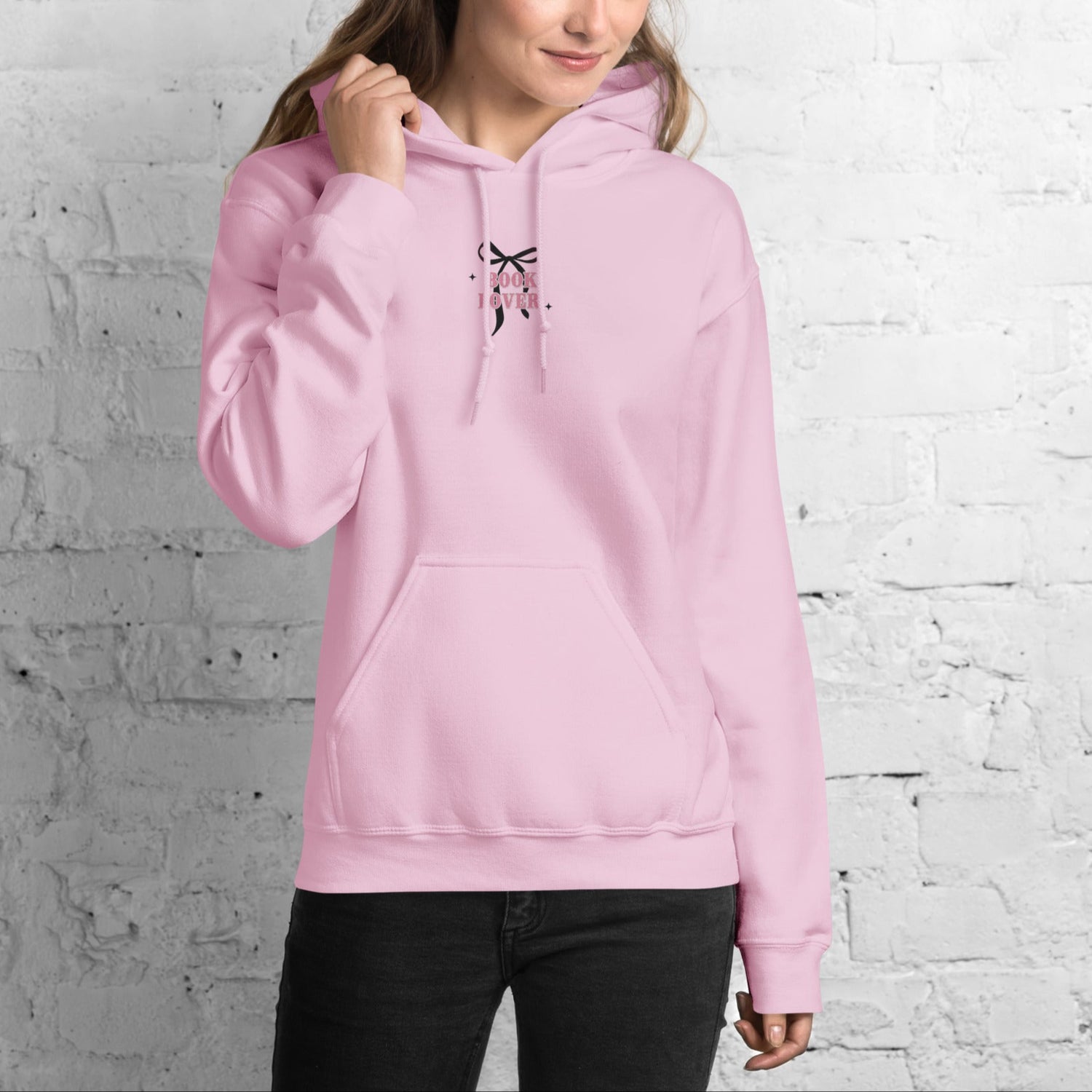 Woman wearing a pink book lover hoodie with an embroidered bow against a white brick wall