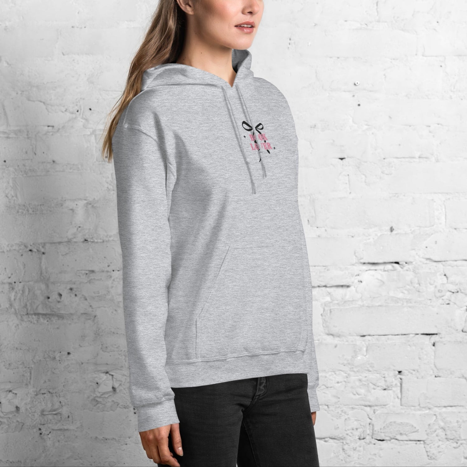 Woman wearing a gray book lover hoodie with an embroidered bow against a white brick wall