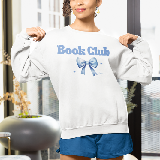Person wearing a white sweatshirt with 'Book Club' and a blue bow graphic, standing indoors.